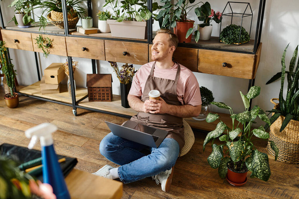 Mężczyzna spokojnie siedzi na podłodze, kołysząc filiżankę kawy w przytulnym sklepie z roślinami. - Zdjęcie, obraz