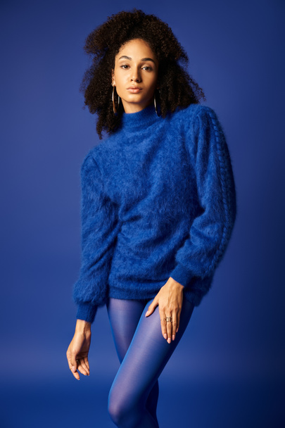Μια νεαρή γυναίκα ποζάρει με χάρη σε ένα ζωηρό μπλε πουλόβερ και καλσόν σε ένα φόντο που ταιριάζει σε ένα σκηνικό στούντιο. - Φωτογραφία, εικόνα