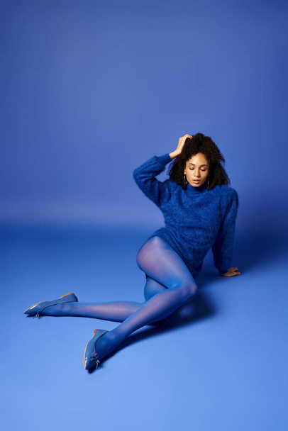 Genç bir kadın zarif bir şekilde yerde oturuyor, bacakları çapraz, parlak taytlar ve mavi bir arka planda süveter giyiyor.. - Fotoğraf, Görsel