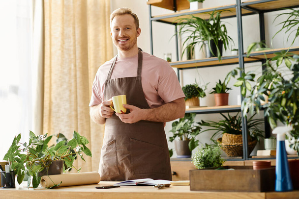 Um homem bonito em um avental desfruta de uma xícara de café em uma loja de plantas, incorporando a essência de possuir uma pequena empresa. - Foto, Imagem