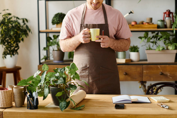 Een man in een schort die geniet van een kopje koffie in een plantenwinkel, een moment rust neemt en geniet van de warme drank. - Foto, afbeelding