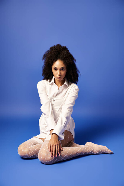 Egy fehér ruhás fiatal nő ül a földön keresztbe tett lábakkal, békességet és eleganciát sugározva egy stúdióban.. - Fotó, kép