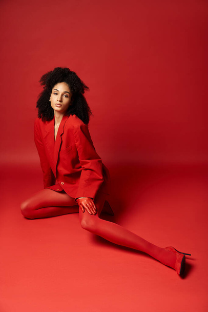 Молодая женщина изысканно одета в красный костюм и колготки, изящно сидящая на ярком полу студии. - Фото, изображение