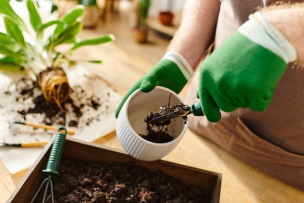 Ένα άτομο με πράσινα γάντια που χύνει χώμα σε ένα δοχείο σε ένα φυτώριο, ενσωματώνοντας την ιδέα της ιδιοκτησίας μιας μικρής επιχείρησης. - Φωτογραφία, εικόνα