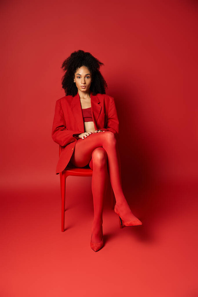 Młoda kobieta w czerwonej kurtce i rajtuzach siedzi elegancko na tętniącym życiem krześle ze skrzyżowanymi nogami. - Zdjęcie, obraz