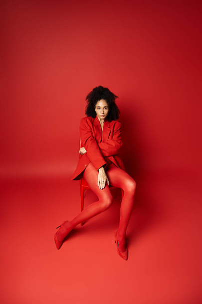 Kırmızı ceketli ve taytlı genç bir kadın canlı kırmızı bir odada, bir sandalyede huzur içinde oturuyor.. - Fotoğraf, Görsel