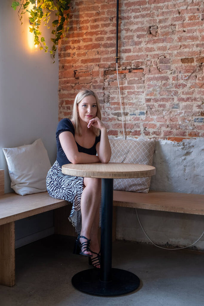 This image portrays a young Caucasian woman with blonde hair seated at a table in a chic cafe, her reflective gaze suggesting deep thought or contemplation. The warm, ambient light and the trailing - Photo, Image