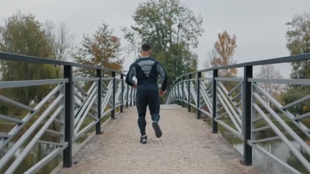 Hombre activo corriendo en un puente de metal en un parque con árboles de otoño. Atleta haciendo entrenamiento cardiovascular. Ejercicios de Jog Actividades. Concepto de estilo de vida saludable y personas - Imágenes, Vídeo