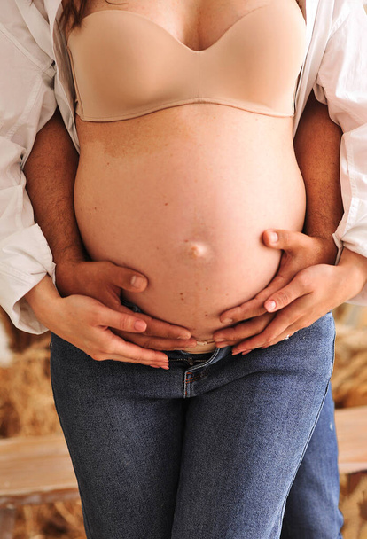 Крупним планом фото вагітної жінки в білій сорочці і блакитних джинсах, а її чоловік тримає руки на вагітному животі. Любовний концепт. Сімейна концепція. Поняття щасливого батьківства - Фото, зображення