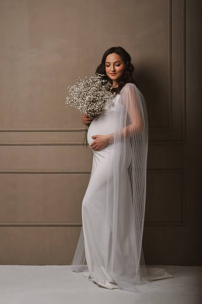 Kaunis raskaana oleva nainen valkoisessa mekossa, jolla on kimppu mustalaisia toisessa kädessä ja hänen raskaana oleva vatsa toisessa kädessä. Heilutti hiuksiaan. Onnellisen äitiyden käsite. Kopioi tilaa tekstille - Valokuva, kuva