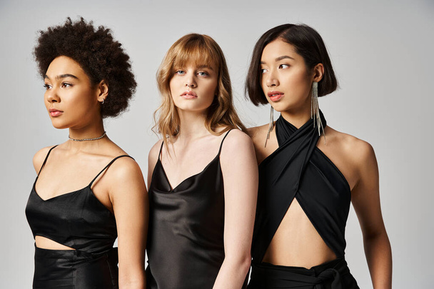 Три жінки різних національностей витончено стоять у чорних сукнях на фоні нейтральної сірої студії. - Фото, зображення