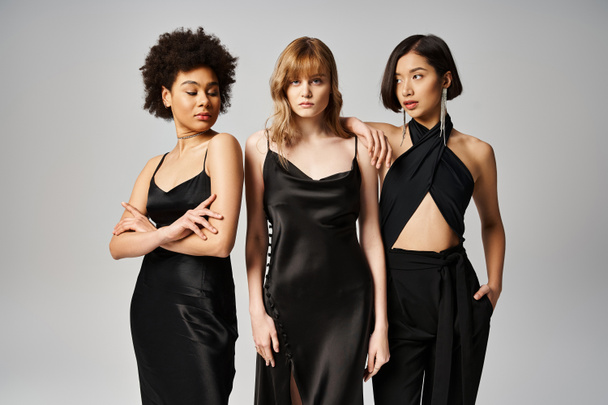 Tres mujeres, cada una con un elegante vestido negro, se paran elegantemente juntas sobre un fondo gris de estudio. - Foto, imagen