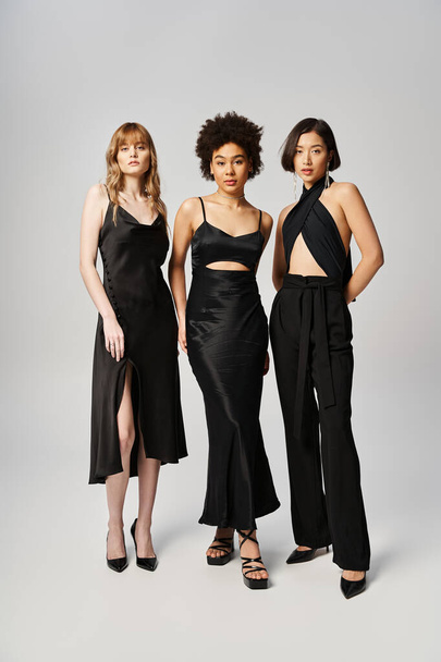Drei Frauen verschiedener Ethnien stehen in schwarzen Kleidern in einem Studio vor grauem Hintergrund zusammen. - Foto, Bild