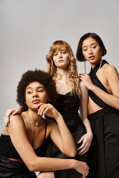 Τρεις γυναίκες με διαφορετικό υπόβαθρο σε μαύρα φορέματα θέτουν με χάρη σε ένα στούντιο σε ένα γκρι φόντο. - Φωτογραφία, εικόνα