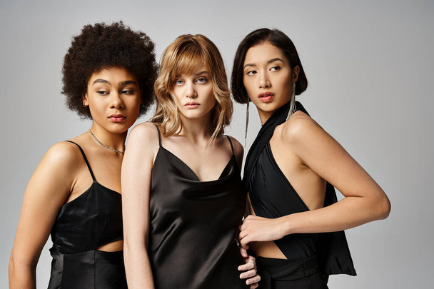 Τρεις γυναίκες με μαύρα φορέματα ποζάρουν για μια φωτογραφία σε ένα γκρίζο φόντο στούντιο, αναδεικνύοντας την ομορφιά και την ποικιλομορφία. - Φωτογραφία, εικόνα