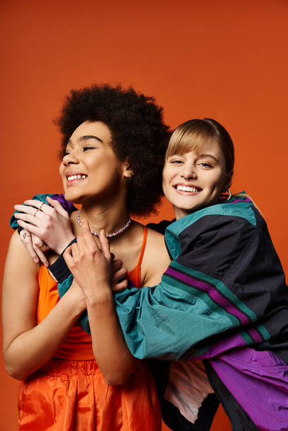 Due donne, di etnie diverse, si chiudono in un caldo abbraccio, sorridono raggianti su uno sfondo arancione. - Foto, immagini