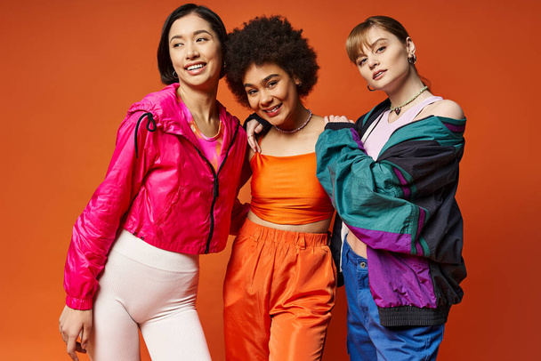 Drie vrouwen van verschillende etniciteiten staan vol vertrouwen in een studio met een oranje achtergrond, die multiculturele schoonheid etaleert. - Foto, afbeelding