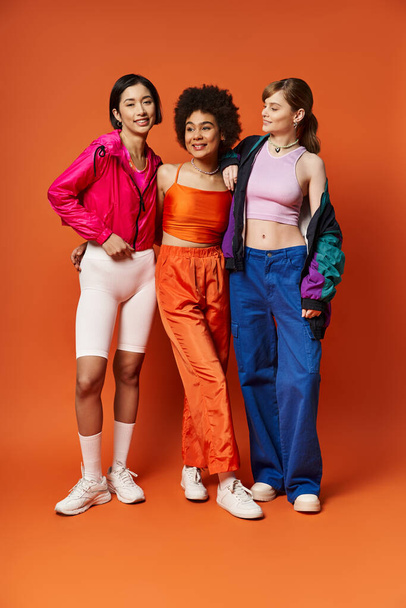 Three women of diverse backgrounds stand united, representing beauty and strength against an orange studio backdrop. - Foto, Bild