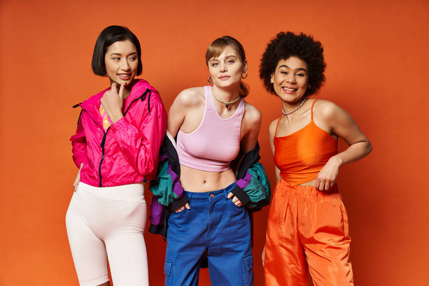 Três mulheres de diferentes etnias em pé juntas, representando diversidade e unidade em um fundo laranja vibrante. - Foto, Imagem