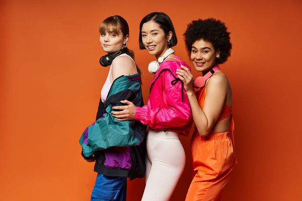 Três mulheres de diferentes etnias e estilos de pé juntos em um estúdio com um fundo laranja vibrante. - Foto, Imagem