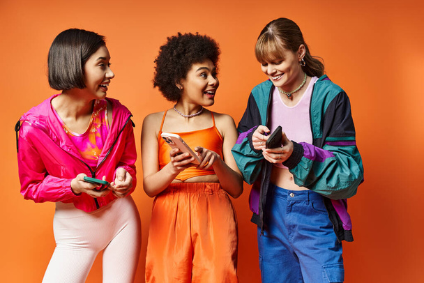 Tre giovani donne diverse ridono e guardano i loro telefoni cellulari su un vibrante sfondo arancione. - Foto, immagini