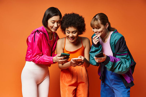Drie verschillende meisjes, waaronder blanke, Aziatische en Afro-Amerikaanse, huddled samen op zoek naar een mobiele telefoon met een oranje achtergrond. - Foto, afbeelding