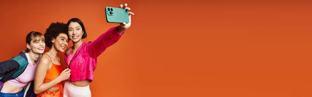 Multicultural women taking a selfie on a cell phone against an orange studio background in a joyful moment. - Foto, Bild