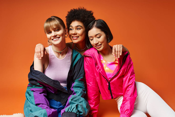 Üç farklı kadın zarif bir şekilde yerde oturuyor, canlı turuncu bir arka plan tarafından çerçevelenmiş bir resim için gülümsüyor.. - Fotoğraf, Görsel