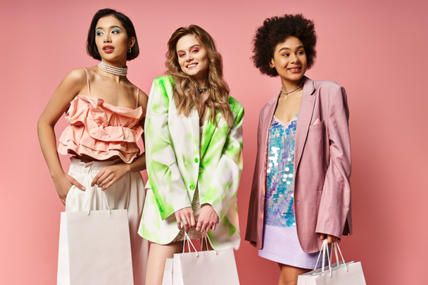 Drei Frauen unterschiedlicher Herkunft stehen zusammen und halten Einkaufstüten vor einem leuchtend rosafarbenen Hintergrund. - Foto, Bild