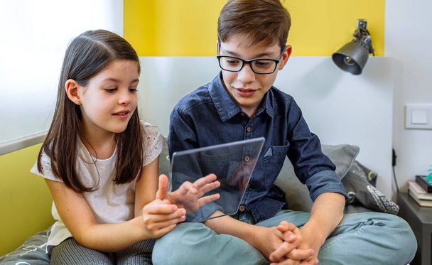 Szczęśliwe dzieci bawiące się futurystyczną przezroczystą tabletką siedzącą nad łóżkiem. Wyświetlacz cyfrowy z koncepcją szklanego ekranu. Uśmiechnięte dzieci korzystające z przenośnego urządzenia w wolnym czasie w domu. - Zdjęcie, obraz