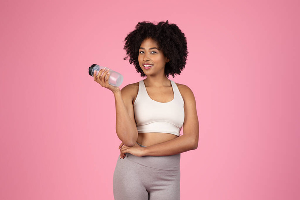 Веселая молодая черная женщина с вьющимися волосами, одета в белый спортивный бюстгальтер и серые леггинсы, держа бутылку воды, с уверенной позой на розовом фоне - Фото, изображение