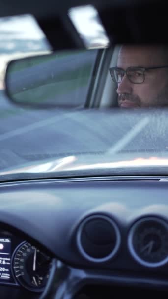 Arabanın dikiz aynasında gözlüklü bir adam. Sürücü yola dikkat ediyor, ön camda ilerleyen yolu görüyoruz, sürüş ve seyahat videosu.. - Video, Çekim