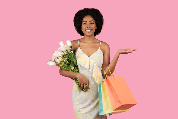 Radostně se usmívající tisíciletá Afroameričanka radostně drží v ruce kytici bílých tulipánů a barevných nákupních tašek, s otevřenou rukou, na růžovém pozadí - Fotografie, Obrázek