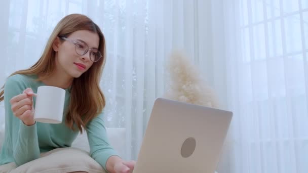 Νεαρή Ασιάτισσα γυναίκα που φοράει γυαλιά καθιστή καναπέ που εργάζεται σε φορητό υπολογιστή και πίνει καφέ στο σαλόνι στο σπίτι, γυναίκα που εργάζεται από το σπίτι με τηλεργασία, ανεξάρτητη εργασία, επιχειρηματική και ψηφιακή ζωή. - Πλάνα, βίντεο