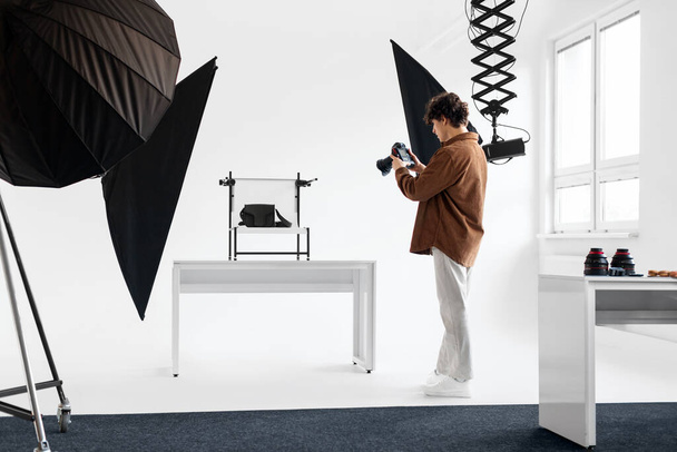 Skoncentrowany fotograf płci męskiej w stroju casual strzela profesjonalne zdjęcie produktu w dobrze oświetlonym studio ustawienie z softbox i sprzętu - Zdjęcie, obraz