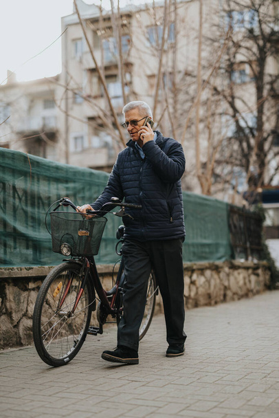 Yaşlı bir beyefendi kentin kaldırımında duruyor, yanında bir bisikletle telefon görüşmesi yapıyor. Sahne, günlük şehir hayatını ve aktif yaşlıları yansıtıyor.. - Fotoğraf, Görsel