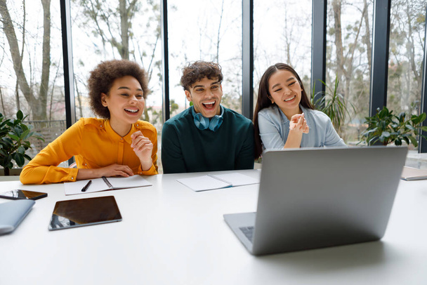 Три веселих різноманітних студента в повсякденному одязі захоплені і сміються, коли вони дивляться на екран ноутбука в яскравому, сучасному навчальному просторі з видом на вікно - Фото, зображення