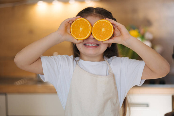 Retrato de adorable niña en delantal de pie en la cocina y sonriendo. KId sosteniendo mitades de naranjas en sus ojos. .. Foto de alta calidad - Foto, imagen