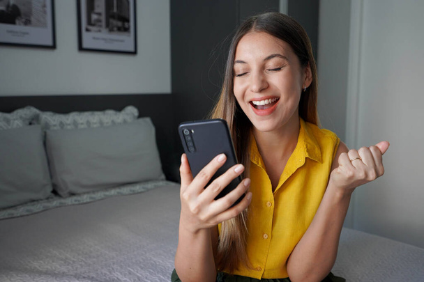 Mujer brasileña joven emocionada por el éxito sosteniendo el teléfono inteligente con el puño en alto y los ojos cerrados celebrando la victoria sonriendo en su habitación en casa. Concepto ganador. - Foto, imagen