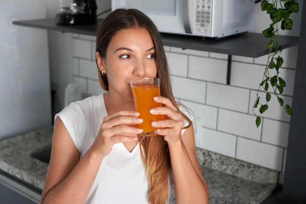 Dziewczyna pijąca sok mandarynkowy w kuchni. Młoda kobieta pije rano w domu sok pomarańczowy lub marchewkowy. - Zdjęcie, obraz