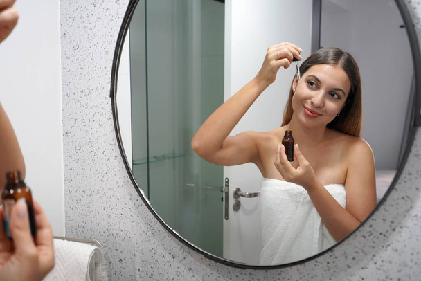 Όμορφη νεαρή γυναίκα που εφαρμόζει υαλουρονικό οξύ με πιπέττα στο μπάνιο - Φωτογραφία, εικόνα
