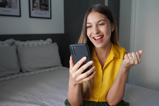 Felice giovane donna brasiliana in possesso di smartphone e celebra con un pugno serrato seduto sul letto a casa - Foto, immagini
