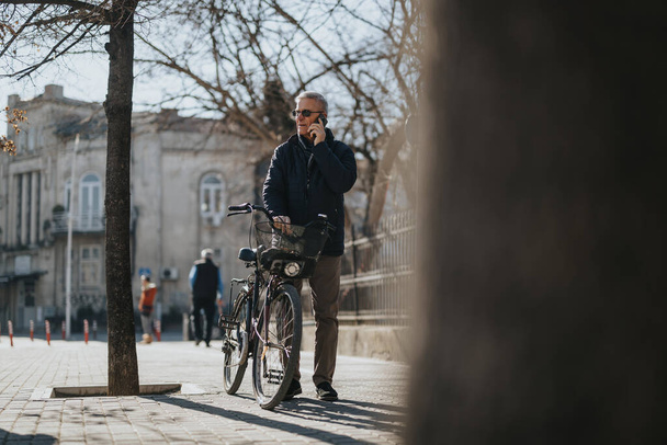 Egy középkorú férfi alkalmi ruhában telefonbeszélgetést folytat, biciklijét egy városi utcán tartja egy fényes, napsütéses napon.. - Fotó, kép