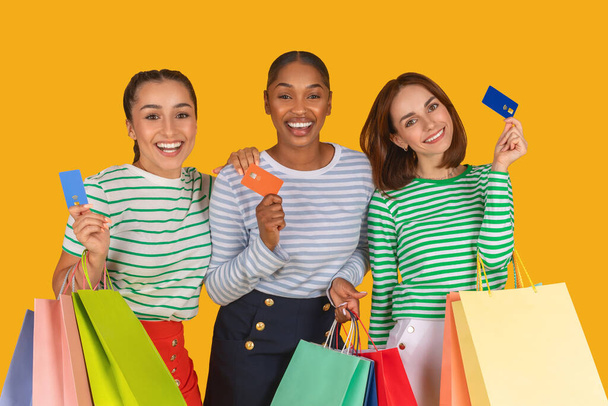Sales And Discounts Concept. Smiling excited multiracial women holding credit card and shopping bags, got cashback, looking at camera, standing isolated over bright yellow studio background - Photo, Image