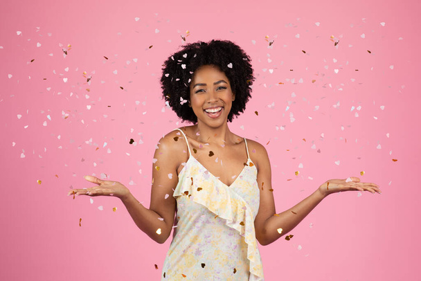 Binlerce yıllık parlak gülümseyen kıvırcık saçlı Afro-Amerikan kadın konfeti yağmurunun ortasında neşeyle gülüyor, tatilin tadını çıkarıyor, canlı pembe bir arka planda, stüdyoda. Eğlence, etkinlik - Fotoğraf, Görsel
