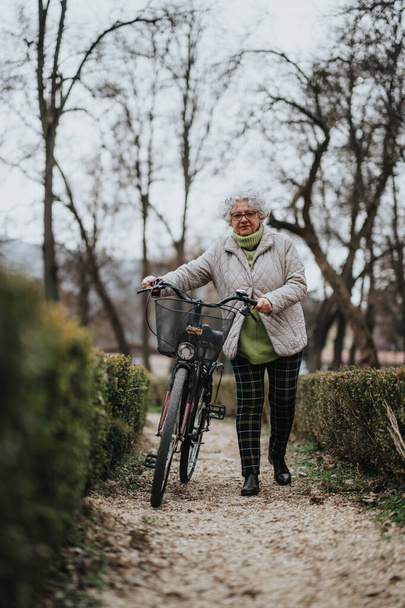 Volwassen vrouwelijke gepensioneerde wandelen met een fiets door een rustig park, belichaamt een actieve levensstijl en onafhankelijkheid. - Foto, afbeelding