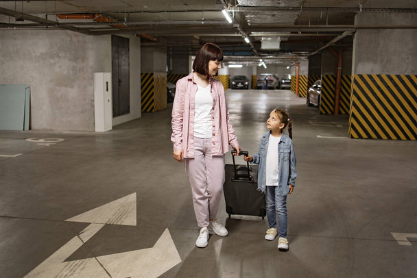 Happy mom and small daughter, attired in informal wear, stroll through subterranean parking area. Youthful lady with her child clutching travel suitcase searching for vehicle before starting journey. - Foto, afbeelding