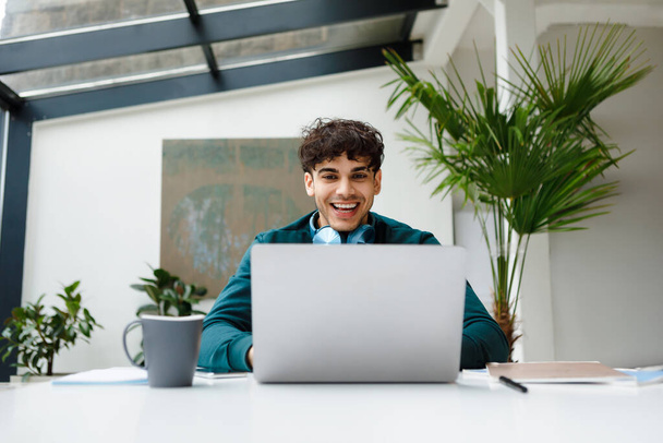 Radosny młody freelancer z modnymi słuchawkami śmieje się podczas angażowania się w treści na laptopie w słonecznym, wypełnionym roślinami miejscu pracy, pokazując radość - Zdjęcie, obraz