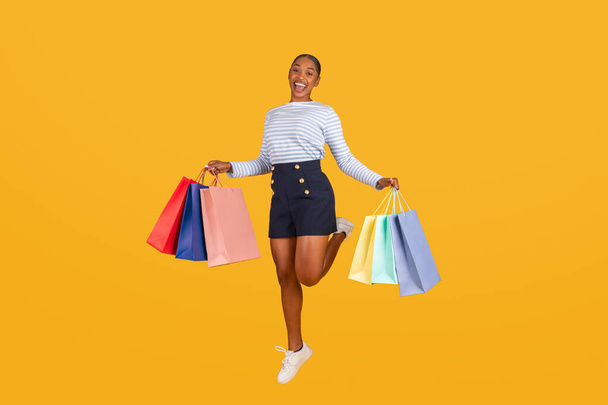 Щаслива молода афроамериканка стрибає в повітрі з сумками в руках ізольовано на жовтому студійному фоні. Радісна чорна леді в літньому одязі насолоджується продажем сезону - Фото, зображення