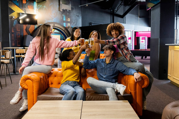 Cheerful friends toasting with beverages at a colorful arcade lounge. - Photo, Image